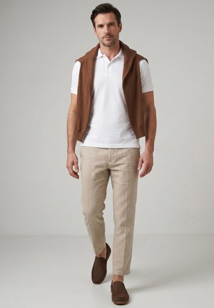 Mann, der geht, trägt ein weißes Polohemd, einen braunen Pullover über den Schultern, beige Hosen und braune Loafers vor einfarbigem Hintergrund.