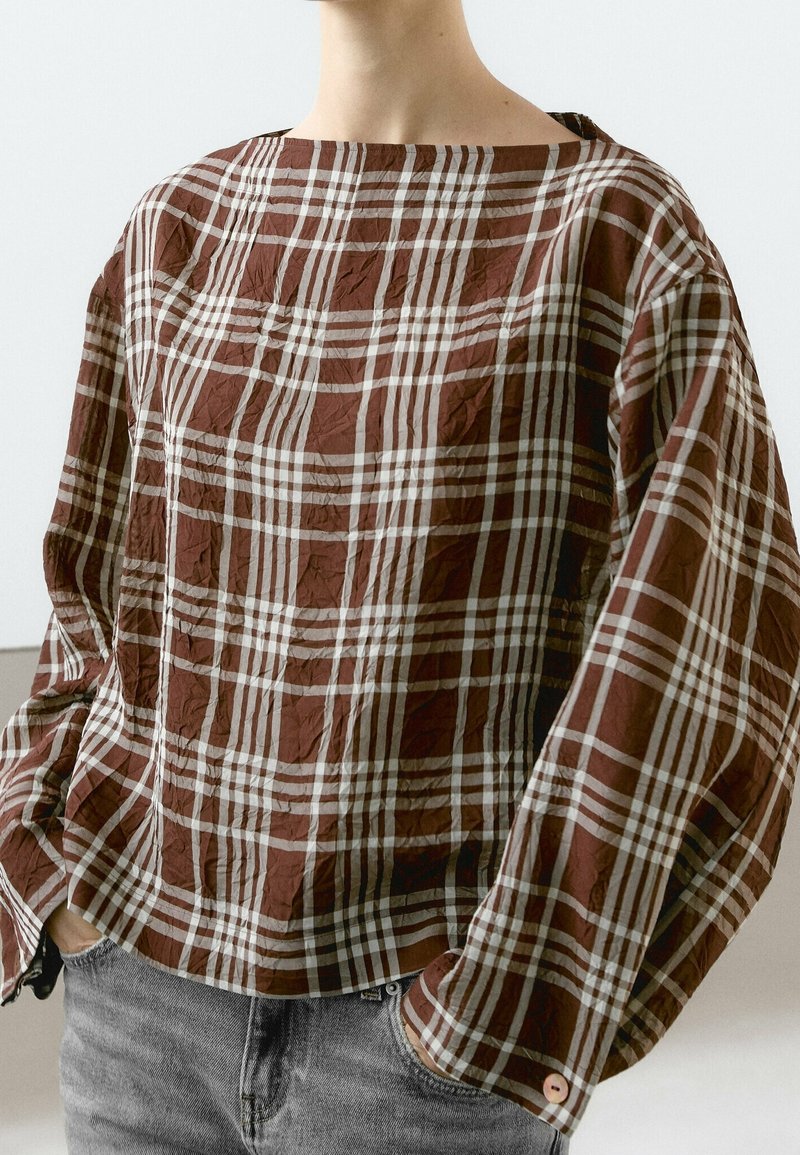 Personne portant une chemise ample à carreaux marron et blancs à manches longues, les mains dans les poches d’un jean gris clair, coupée au niveau du cou.