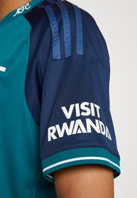 Maglietta sportiva a maniche corte in teal e blu navy con accenti a strisce blu. Presenta "VISIT RWANDA" in bianco sulla manica sinistra.