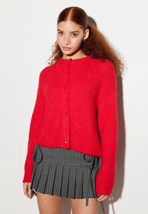 Rode gebreide cardigan met een ronde halslijn, lange mouwen en knoopsluiting. Draagt over een grijze plisségoden rok met strepen en accentdetails.