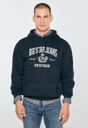 Młody mężczyzna o krótkich włosach, ubrany w ciemnoniebieską bluzę z kapturem Big Star Jeans Heritage na koszuli w kratę i niebieskich dżinsach, stojący na jednolitym tle.