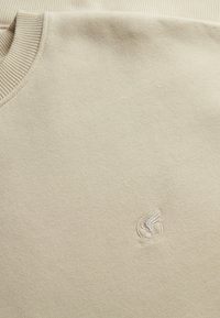 Beżowa bluza z okrągłym dekoltem, prążkowanym kołnierzem i małym, haftowanym logo w kształcie skrzydeł na piersi, wykonana z miękkiego materiału.