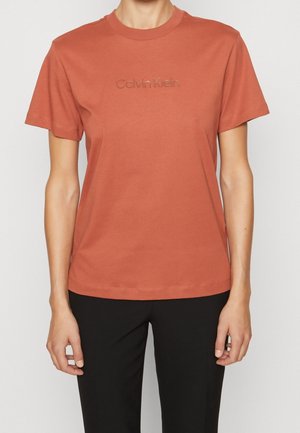 Osoba ubrana w krótki, rdzawo-brązowy t-shirt Calvin Klein oraz czarne spodnie, pokazana od szyi do bioder, zwrócona przodem.