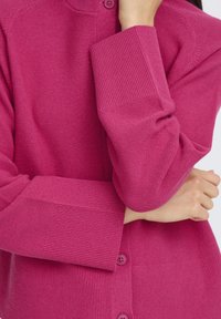 Cardigan tricotat de culoare fucsia, cu textură similară cu ribele, închidere prin butoni pe față și mâneci lungi. Prezintă un gât rotund și o finisare netedă.