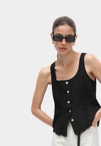 Zwarte linnen mouwloze top met een vierkante halslijn, vijf witte knopen en een rafelige zoom. Model draagt zwarte vierkante zonnebril.