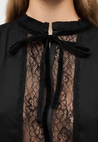 Κοντινό πλάνο μιας μαύρης μπλούζας με διαφανές μπροστινό μέρος από δαντέλα με λουλούδια και βελούδινο φιόγκο δεμένο στον λαιμό.