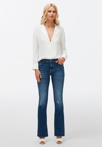 Chemisier blanc à boutons avec un décolleté plongeant, associé à un jean bleu évasé avec des déchirures et des escarpins blancs à bout ouvert.