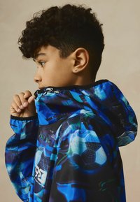 Giacca multicolore con motivi geometrici blu e neri, cappuccio, chiusura con zip e polsini elastici. Materiale leggero, vestibilità sportiva.