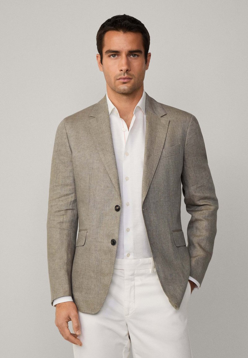 Blazer en lin de couleur gris clair avec deux boutons, revers crantés et deux poches avant, associé à une chemise blanche et un pantalon clair.