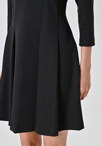 Schwarzes Kleid mit tailliertem Oberteil und auslaufendem Rock. Hergestellt aus strukturiertem Stoff, mit horizontalen Falten und dreiviertellangen Ärmeln.