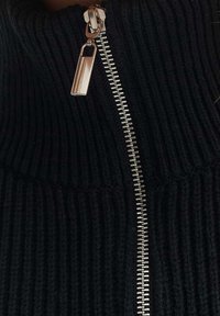 Fermeture éclair argentée partiellement fermée sur un tissu en maille côtelée noire, vue rapprochée du curseur et de la texture du textile.