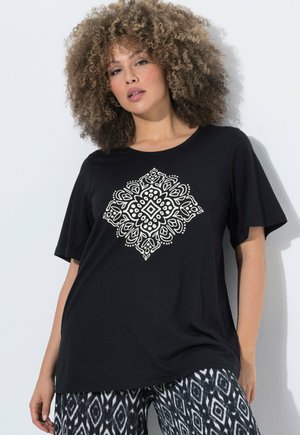 Frau mit lockigem Haar trägt ein schwarzes T-Shirt mit weißem symmetrischem Muster und schwarz-weißer Hose mit geometrischem Druck, steht vor einer weißen Wand.