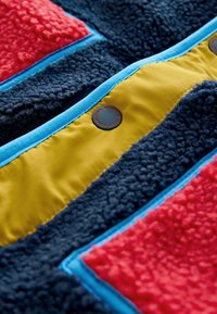 Țesătură de fleece multicolor cu secțiuni roșii, bleumarin, galbene și albastre, având o suprafață texturată și un detaliu cu un buton circular negru.