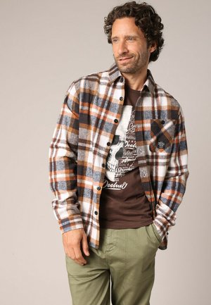 Chemise en flanelle à motif à carreaux orange, bleu et blanc, avec un devant à boutons et une poche sur la poitrine. Portée par-dessus un t-shirt graphique marron.
