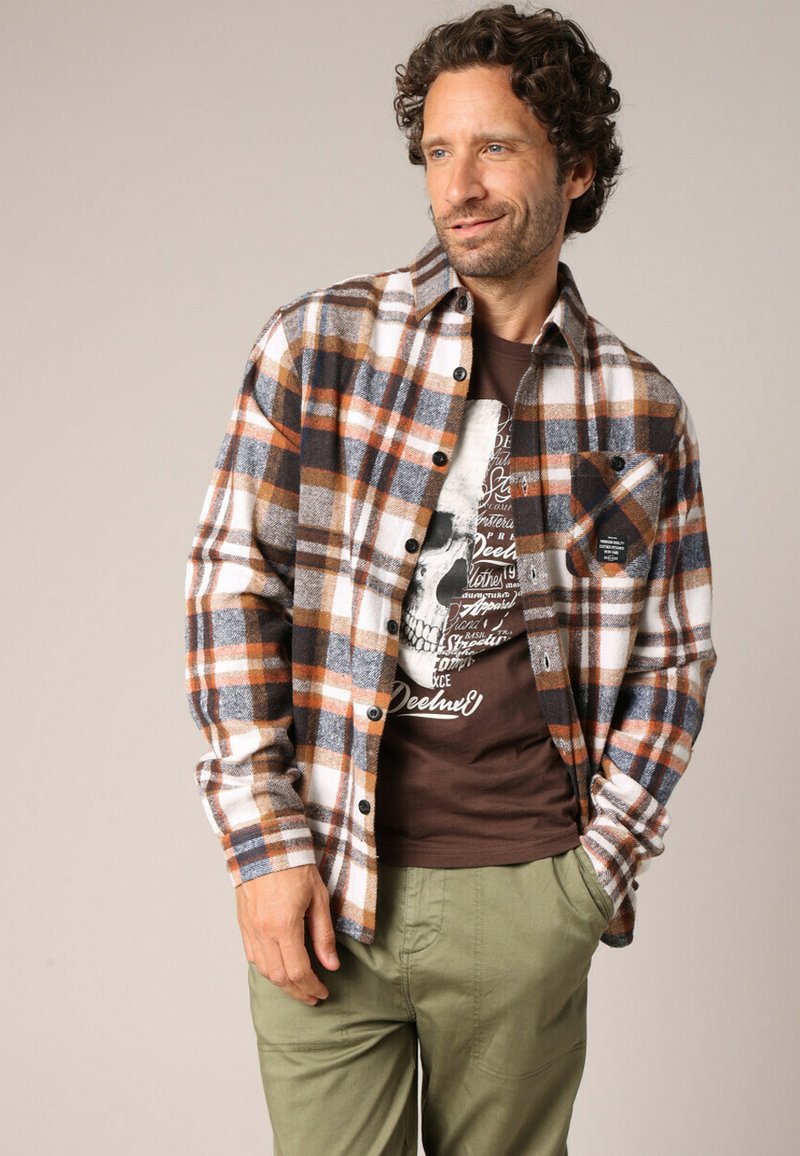 Flanellen shirt in oranje, blauw en witte geruite patroon met een knoopsluiting aan de voorkant en een borstzak. Gemaakt over een bruine graphic t-shirt.