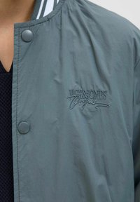 Ljusgrå jacka med en slät textur, tryckknappsstängning och vitrandig krage; har broderad "Jack & Jones"-logotyp.