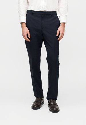 Mand iført sorte skræddersyede bukser, brune pæne sko og en hvid langærmet skjorte med knapper, stående mod en hvid baggrund.