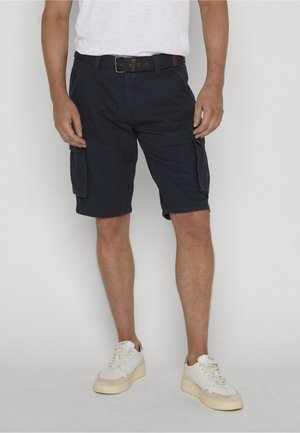Mand iført sorte cargoshorts med sidelommer, sort bælte, hvide sneakers og en hvid T-shirt, stående mod en ensfarvet baggrund.