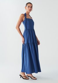 Modré maxi šaty s priliehavým živôtikom, širokými ramienkami s kruhovými ozdobami a viacvrstvovou sukňou. Vyrobené z ľahkej látky s hladkým povrchom.