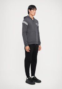 Szürke cipzáras kapucnis pulóver csíkos vállbetétekkel, fekete joggerekrel és fekete sneakers-szel párosítva. Simább anyag és laza stílus.