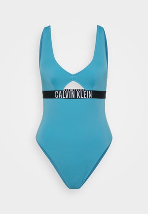 Bañador de una pieza color turquesa con escote en V profundo, detalle recortado y una banda negra con el logotipo de Calvin Klein bajo el busto. Textura de tela suave.
