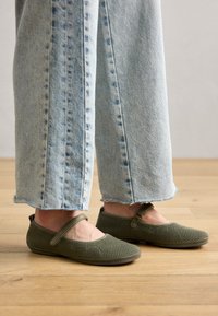 Zapatos planos Mary Jane tejidos de color verde con una correa, punta redonda y parte superior texturizada, combinados con unos jeans de tiro bajo y dobladillo deshilachado de color azul claro, sobre un suelo de madera.