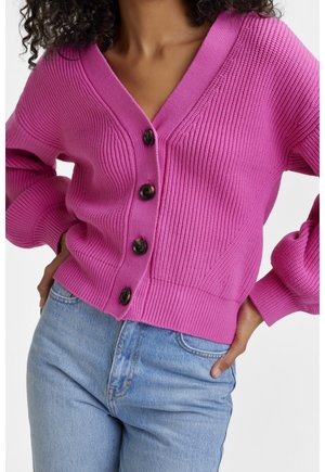 Kobieta w jaskraworóżowym prążkowanym kardiganie z czterema czarnymi guzikami i jasnoniebieskich wysokich dżinsach, z rękami opartymi na biodrach.