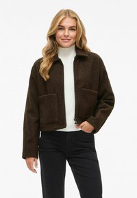 Veste brune, douce et courte avec une fermeture éclair sur le devant, deux poches avant et un col, portée sur une chemise blanche et associée à un pantalon noir.