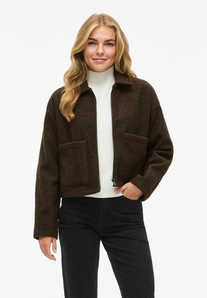 Veste brune, douce et courte avec une fermeture éclair sur le devant, deux poches avant et un col, portée sur une chemise blanche et associée à un pantalon noir.