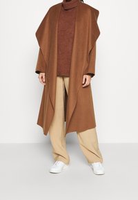 Brązowy, oversize'owy wełniany płaszcz z szerokimi klapami spoczywa na rudym golfie, zestawiony z luźnymi beżowymi spodniami i białymi tenisówkami.