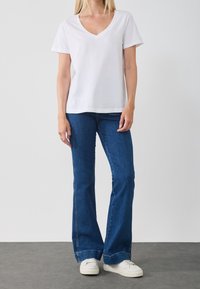 En person klädd i en vit V-ringad kortärmad t-shirt, blå utsvängda jeans och vita sneakers står mot en enkel vägg.