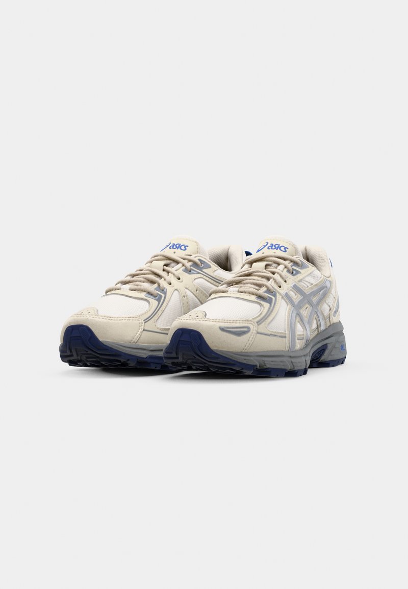 Zapatillas de running beige con parte superior texturizada, acentos en azul y suela de goma duradera; presenta un logo en la lengüeta y sobrepuestos en los lados.