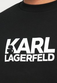 Čierne bavlnené tričko s okrúhlym výstrihom, na ktorom je výrazný biely nápis: "KARL" nad slovom "LAGERFELD" a siluetou muža.