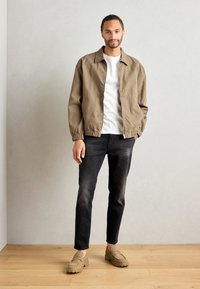 Jachetă bomber maro deschis cu manșete elastice, tricou alb, blugi negri decolorati și pantofi bej cu talpă groasă. O ținută simplă și lejeră.