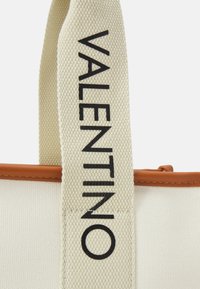 Sac fourre-tout en toile avec un corps beige, des accents en cuir marron et un logo noir "VALENTINO" bien visible sur la poignée.