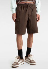 Bruna cargoshorts med elastisk midja och dragsko, med sidoficka, bärs med svarta strumpor och bruna sneakers.