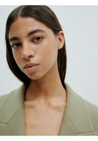 Close-upportret van een persoon met steil, donkerbruin haar, die een lichtgroene blazer met gestructureerde schouders en een diepe halslijn draagt.