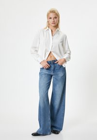 Biała koszula zapinana na guziki z długimi rękawami, zestawiona z jasnoniebieskimi spodniami typu wide-leg. Modelka stoi z rękami na biodrach, na minimalistycznym tle.