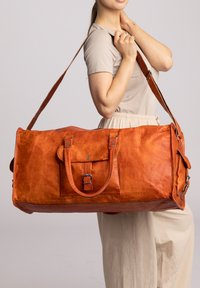 Mittelgroße orangefarbene Leder-Duffel-Tasche mit doppelten Griffen, verstellbarem Schulterriemen und Fronttasche mit Schnallenverschluss. Glatte Textur.