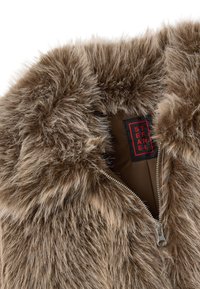 Collar de chaqueta de piel sintética marrón con una cremallera parcialmente abierta y una etiqueta negra con texto rojo en su interior.