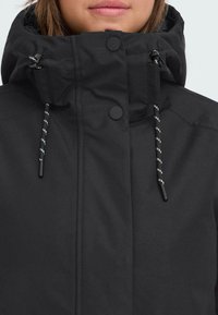 Svart utomhusjacka med hög krage, framknäppning och dragsko med vita detaljer. Tillverkad av ett slätt och hållbart material.