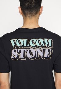 Personne aux cheveux courts et bouclés portant des boucles d'oreilles créoles en or et une chemise noire avec un grand texte "Volcom Stone" en bleu et violet au dos.