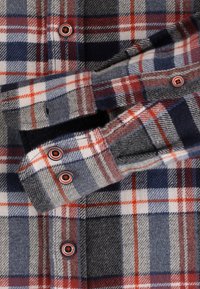 Geruite flanellen overhemd met een rood, marineblauw, grijs en wit ruitpatroon, voorzien van donkere knopen met rode en witte concentrische ringen.