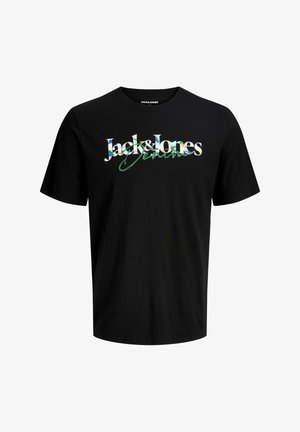 Ξανθό αγόρι που χαμογελά, φοράει μαύρο T-shirt Jack & Jones, άνετα μπλε τζιν και μπεζ αθλητικά παπούτσια, στέκεται μπροστά σε απλό φωτεινό φόντο.