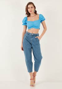 Top corto blu con maniche a sbuffo e parte anteriore arricciata, abbinato a jeans in denim chiaro a vita alta. Il modello indossa sandali con straps multicolori.