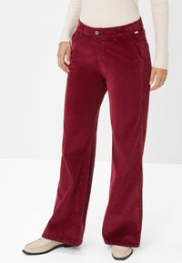 Rote Cord-Hosen mit weitem Bein und strukturiertem Finish, ausgestattet mit Fronttaschen und einem Knopfverschluss. Kombiniert mit einem enganliegenden cremefarbenen Oberteil.