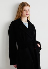 Μαύρο μάλλινο παλτό με ζώνη στη μέση, μεγάλη τσέπη και φαρδιές πατιλέτες. Διαθέτει άνετη εφαρμογή με μακριά μανίκια και λεία υφή.