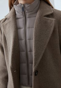 Manteau en laine marron avec une veste intérieure matelassée crème ajustée. Il dispose d'un col haut et d'une fermeture zippée. Tissu texturé avec une doublure lisse.