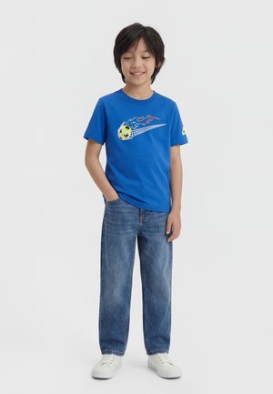 Ragazzo in piedi con una mano in tasca, indossa una t-shirt blu con grafica di pallone da calcio fiammeggiante, jeans blu larghi e sneaker bianche.