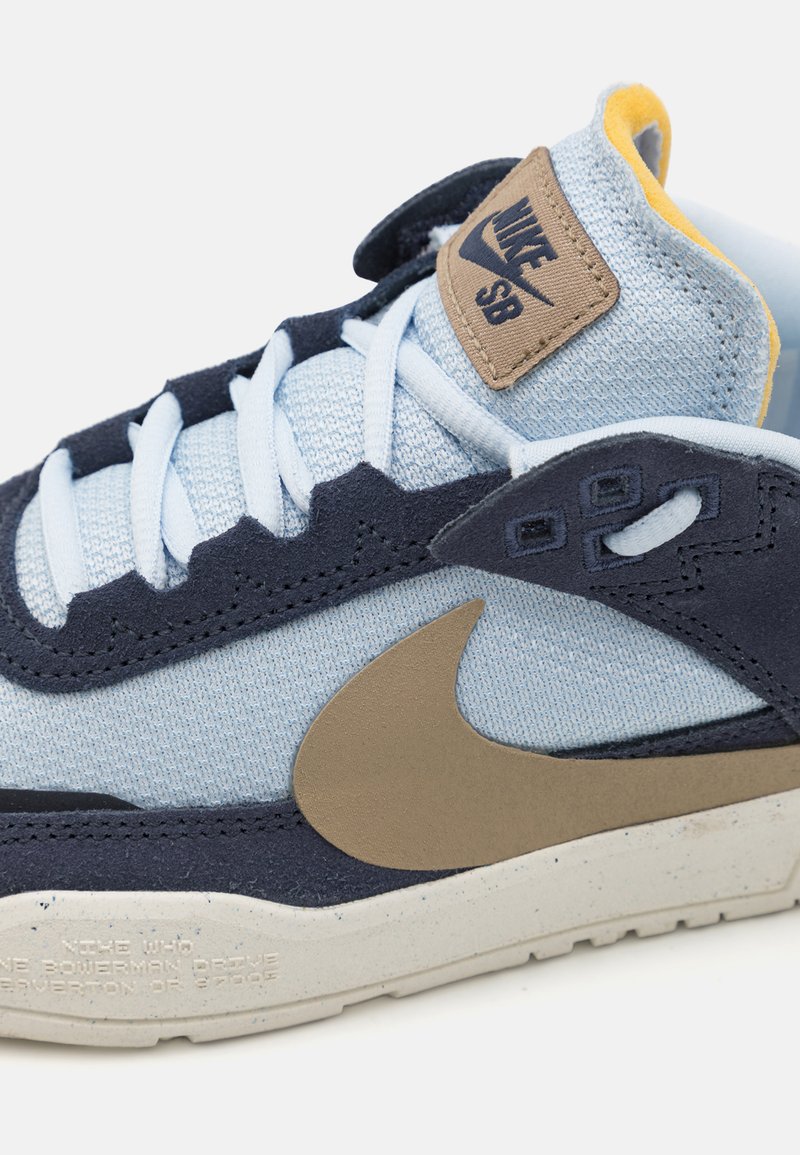 Marineblauer und hellblauer Sneaker aus Textil und Wildleder, mit einem beigen Logo-Patch, geformter Sohle und einem beigen Swoosh-Design.